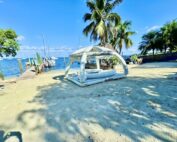 A sunny beach scene at Key Lime Sailing Club in the Florida Keys with a large white AquaBanas inflatable relaxation raft on the sand, surrounded by palm trees and sailboats docked offshore in the clear blue water.
